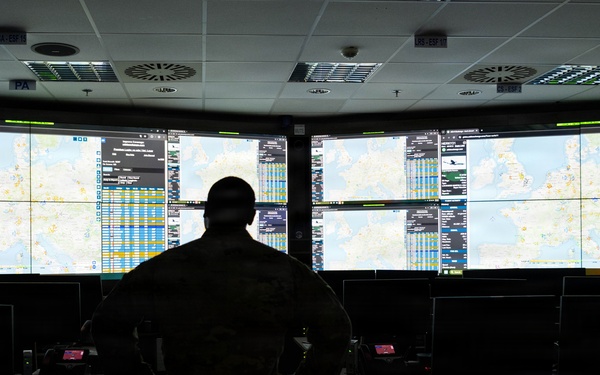 A silhouetted individual in military camouflage uniform stands before a large multi-screen video wall displaying geographic maps and data tables in a darkened operations or command center. The room contains multiple workstations with monitors, keyboards, and equipment arranged in rows. The large display wall appears to show air traffic or operational tracking information over European geography.