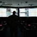 A silhouetted individual in military camouflage uniform stands before a large multi-screen video wall displaying geographic maps and data tables in a darkened operations or command center. The room contains multiple workstations with monitors, keyboards, and equipment arranged in rows. The large display wall appears to show air traffic or operational tracking information over European geography.