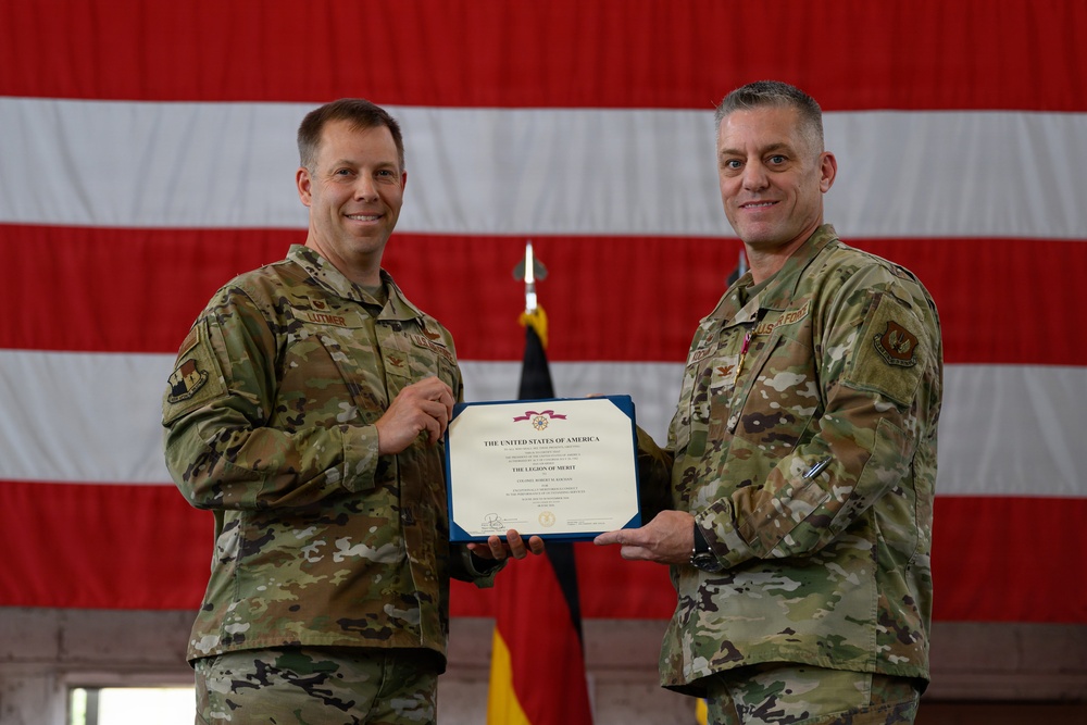 Two U.S. Air Force personnel in OCP (Operational Camouflage Pattern) uniforms are posing together while holding a Legion of Merit certificate issued by the United States of America. The ceremony appears to be an awards presentation taking place in a hangar or large indoor military facility. A large American flag and what appears to be a German flag are visible in the background.