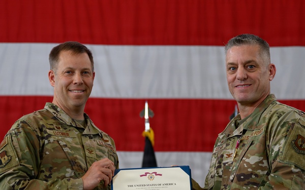 Two U.S. Air Force personnel in OCP (Operational Camouflage Pattern) uniforms are posing together while holding a Legion of Merit certificate issued by the United States of America. The ceremony appears to be an awards presentation taking place in a hangar or large indoor military facility. A large American flag and what appears to be a German flag are visible in the background.