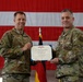 Two U.S. Air Force personnel in OCP (Operational Camouflage Pattern) uniforms are posing together while holding a Legion of Merit certificate issued by the United States of America. The ceremony appears to be an awards presentation taking place in a hangar or large indoor military facility. A large American flag and what appears to be a German flag are visible in the background.