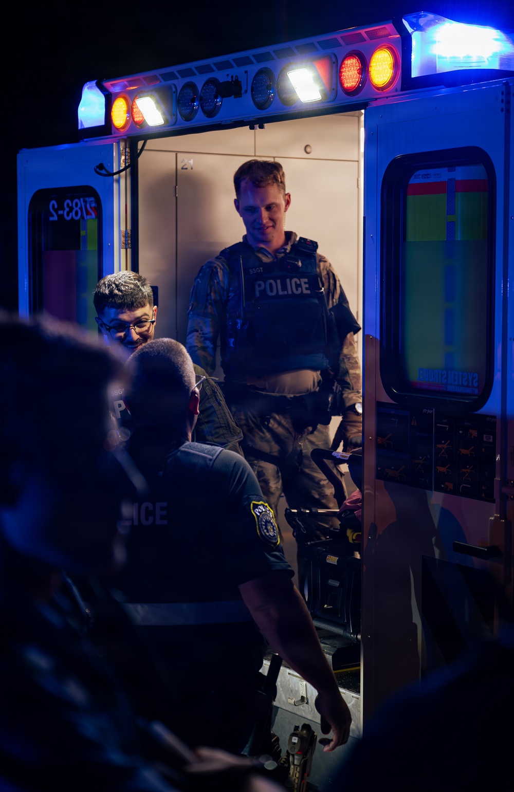 A nighttime scene showing multiple law enforcement personnel gathered at the open rear doors of an ambulance, illuminated by blue emergency lights. A military or tactical police officer wearing a vest labeled 'POLICE' and rank insignia 'SSGT' stands inside the ambulance compartment. Additional officers in police vests are visible in the foreground, appearing to confer or coordinate.