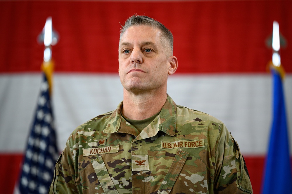 A U.S. Air Force Colonel named Kochan stands at attention in an official military setting. He is wearing Operational Camouflage Pattern (OCP) uniform with visible rank insignia (Colonel's eagle) and name tape. The background features the American flag and what appears to be an Air Force flag, suggesting a formal ceremony or official event.