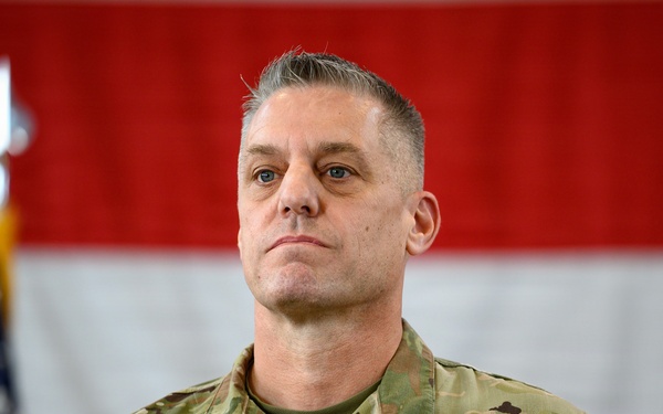 A U.S. Air Force Colonel named Kochan stands at attention in an official military setting. He is wearing Operational Camouflage Pattern (OCP) uniform with visible rank insignia (Colonel's eagle) and name tape. The background features the American flag and what appears to be an Air Force flag, suggesting a formal ceremony or official event.