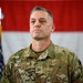A U.S. Air Force Colonel named Kochan stands at attention in an official military setting. He is wearing Operational Camouflage Pattern (OCP) uniform with visible rank insignia (Colonel's eagle) and name tape. The background features the American flag and what appears to be an Air Force flag, suggesting a formal ceremony or official event.