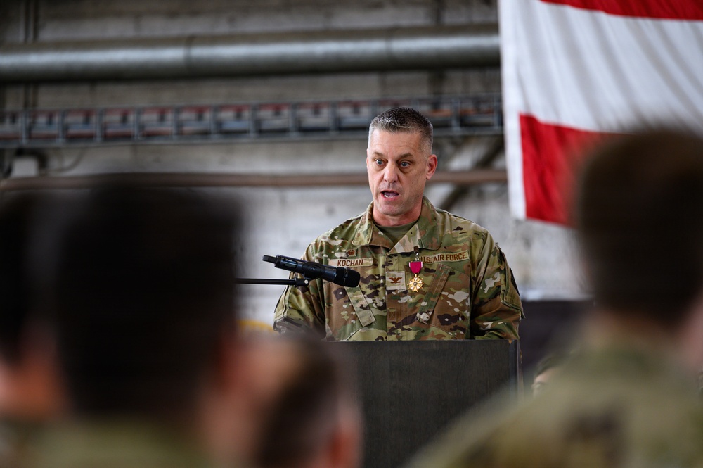 A U.S. Air Force officer with the name tape 'KOCHAN' is speaking at a podium during what appears to be a military ceremony. He is wearing an OCP (Operational Camouflage Pattern) uniform and is decorated with a medal, likely the Meritorious Service Medal or similar award. An American flag is visible in the background, and an audience of uniformed personnel is seated in the foreground.