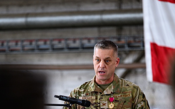 A U.S. Air Force officer with the name tape 'KOCHAN' is speaking at a podium during what appears to be a military ceremony. He is wearing an OCP (Operational Camouflage Pattern) uniform and is decorated with a medal, likely the Meritorious Service Medal or similar award. An American flag is visible in the background, and an audience of uniformed personnel is seated in the foreground.