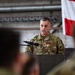 A U.S. Air Force officer with the name tape 'KOCHAN' is speaking at a podium during what appears to be a military ceremony. He is wearing an OCP (Operational Camouflage Pattern) uniform and is decorated with a medal, likely the Meritorious Service Medal or similar award. An American flag is visible in the background, and an audience of uniformed personnel is seated in the foreground.