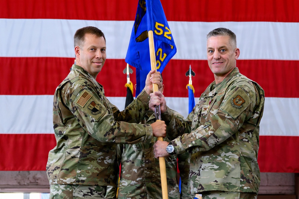 Two U.S. Air Force personnel in OCP (Operational Camouflage Pattern) uniforms are conducting a guidon transfer ceremony, likely a change of command. Both individuals are jointly holding a blue unit guidon bearing the number '52' and an eagle emblem. A large American flag serves as the backdrop, with additional flags visible in the background.