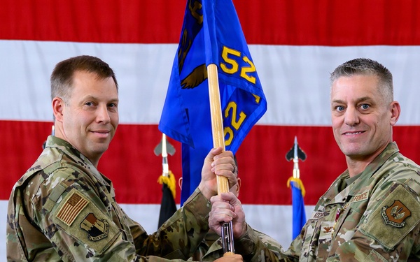 Two U.S. Air Force personnel in OCP (Operational Camouflage Pattern) uniforms are conducting a guidon transfer ceremony, likely a change of command. Both individuals are jointly holding a blue unit guidon bearing the number '52' and an eagle emblem. A large American flag serves as the backdrop, with additional flags visible in the background.