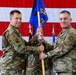 Two U.S. Air Force personnel in OCP (Operational Camouflage Pattern) uniforms are conducting a guidon transfer ceremony, likely a change of command. Both individuals are jointly holding a blue unit guidon bearing the number '52' and an eagle emblem. A large American flag serves as the backdrop, with additional flags visible in the background.