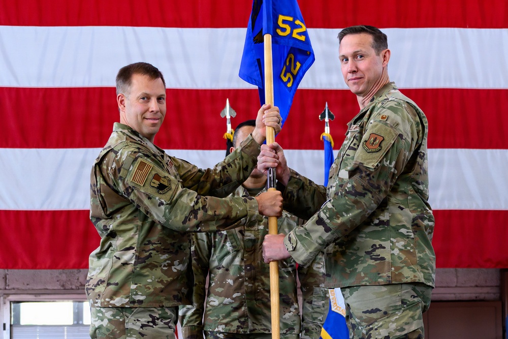 Two U.S. Air Force personnel in OCP (Operational Camouflage Pattern) uniforms are conducting a guidon transfer ceremony, likely a change of command. They are jointly holding a blue military guidon bearing the number '52' in front of a large American flag backdrop. A third uniformed individual is visible in the background holding additional flags.