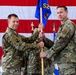 Two U.S. Air Force personnel in OCP (Operational Camouflage Pattern) uniforms are conducting a guidon transfer ceremony, likely a change of command. They are jointly holding a blue military guidon bearing the number '52' in front of a large American flag backdrop. A third uniformed individual is visible in the background holding additional flags.