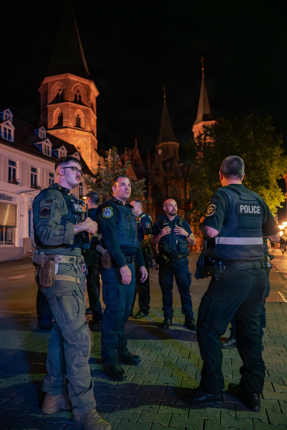 A group of police officers in tactical gear are gathered outdoors at night in what appears to be a European town square or plaza. The officers are wearing dark uniforms with 'POLICE' markings on their vests and appear to be in a briefing or coordination formation. The backdrop features illuminated historic church towers and European-style architecture.