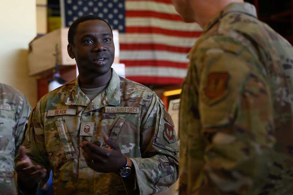 Two U.S. Air Force personnel in OCP (Operational Camouflage Pattern) uniforms are engaged in conversation indoors. The airman facing the camera wears a name tape reading 'BLACK' and displays Master Sergeant rank insignia. An American flag is prominently displayed in the background.