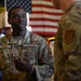 Two U.S. Air Force personnel in OCP (Operational Camouflage Pattern) uniforms are engaged in conversation indoors. The airman facing the camera wears a name tape reading 'BLACK' and displays Master Sergeant rank insignia. An American flag is prominently displayed in the background.