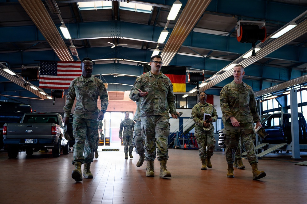 A group of military personnel in MultiCam/OCP camouflage uniforms are walking through a large vehicle maintenance bay or motor pool facility. Two national flags — the United States flag and the German flag — are visibly displayed hanging from the ceiling, suggesting a joint or NATO-affiliated installation. One individual in the center appears to be speaking or briefing the group as they walk.