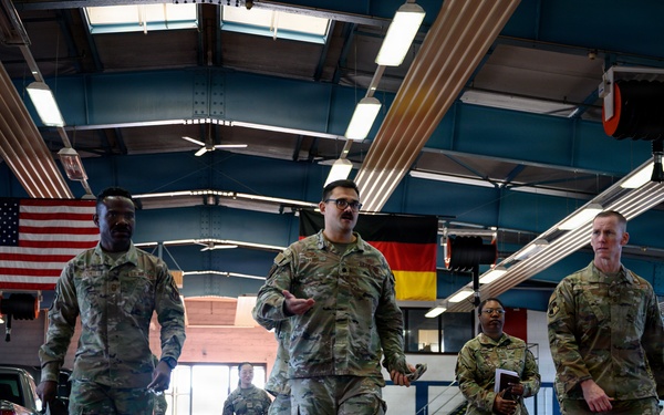 A group of military personnel in MultiCam/OCP camouflage uniforms are walking through a large vehicle maintenance bay or motor pool facility. Two national flags — the United States flag and the German flag — are visibly displayed hanging from the ceiling, suggesting a joint or NATO-affiliated installation. One individual in the center appears to be speaking or briefing the group as they walk.