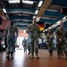 A group of military personnel in MultiCam/OCP camouflage uniforms are walking through a large vehicle maintenance bay or motor pool facility. Two national flags — the United States flag and the German flag — are visibly displayed hanging from the ceiling, suggesting a joint or NATO-affiliated installation. One individual in the center appears to be speaking or briefing the group as they walk.