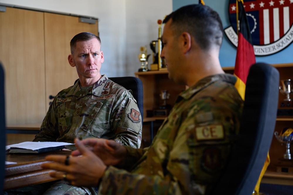 Two U.S. Air Force personnel in OCP (Operational Camouflage Pattern) uniforms are engaged in a meeting or briefing in what appears to be a military office. The individual on the left is attentively listening while the person on the right, wearing a 'CE' (Civil Engineer) tab, appears to be speaking. The office background includes trophies, a German flag, and a unit shield patch on the wall.