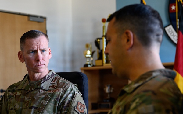 Two U.S. Air Force personnel in OCP (Operational Camouflage Pattern) uniforms are engaged in a meeting or briefing in what appears to be a military office. The individual on the left is attentively listening while the person on the right, wearing a 'CE' (Civil Engineer) tab, appears to be speaking. The office background includes trophies, a German flag, and a unit shield patch on the wall.