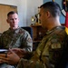 Two U.S. Air Force personnel in OCP (Operational Camouflage Pattern) uniforms are engaged in a meeting or briefing in what appears to be a military office. The individual on the left is attentively listening while the person on the right, wearing a 'CE' (Civil Engineer) tab, appears to be speaking. The office background includes trophies, a German flag, and a unit shield patch on the wall.