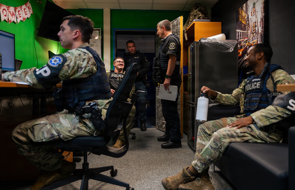A group of law enforcement or military police personnel are gathered in what appears to be a break room or office space. Several individuals wearing camouflage uniforms with 'POLICE' labeled tactical vests are seen relaxing and conversing. The setting has informal decorations including what appears to be Halloween lettering and a New York City Times Square poster.