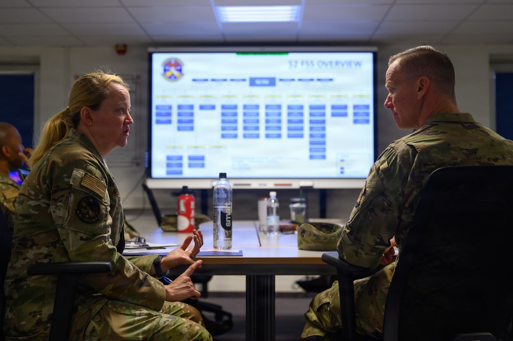Two U.S. military personnel in OCP (Operational Camouflage Pattern) uniforms are seated at a conference table engaged in conversation. A large display screen behind them shows a presentation titled '52 FSS OVERVIEW' with organizational charts and data. The setting appears to be a briefing or operations room at a military installation.