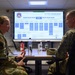Two U.S. military personnel in OCP (Operational Camouflage Pattern) uniforms are seated at a conference table engaged in conversation. A large display screen behind them shows a presentation titled '52 FSS OVERVIEW' with organizational charts and data. The setting appears to be a briefing or operations room at a military installation.