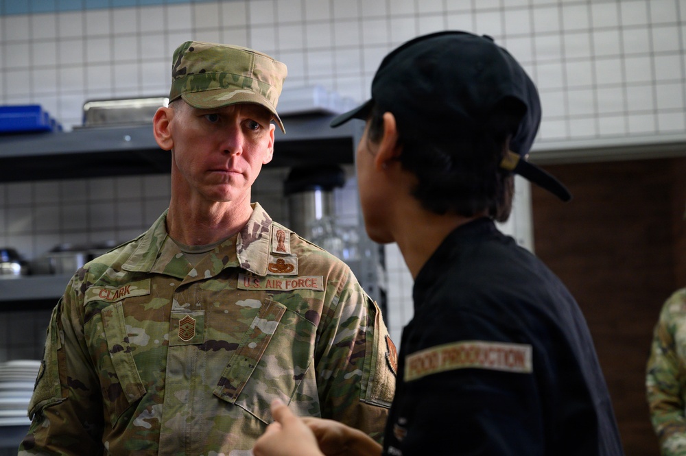 A U.S. Air Force senior enlisted member named Clark, wearing OCP (Operational Camouflage Pattern) uniform, is engaged in conversation with a food service worker in what appears to be a military dining facility kitchen. The airman facing the camera displays a Senior Master Sergeant rank insignia and a U.S. Air Force tape on their uniform. The other individual is wearing a black uniform with a 'Food Production' patch on the back.