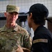A U.S. Air Force senior enlisted member named Clark, wearing OCP (Operational Camouflage Pattern) uniform, is engaged in conversation with a food service worker in what appears to be a military dining facility kitchen. The airman facing the camera displays a Senior Master Sergeant rank insignia and a U.S. Air Force tape on their uniform. The other individual is wearing a black uniform with a 'Food Production' patch on the back.