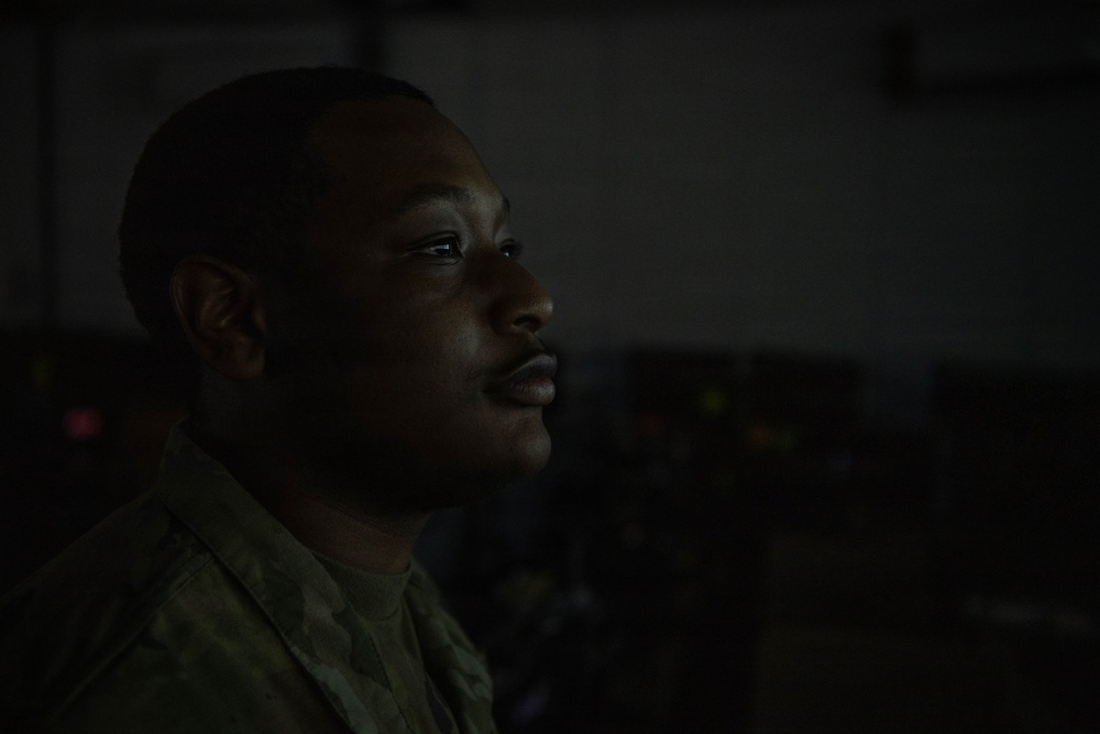 A close-up portrait of a male individual wearing military camouflage uniform, photographed in a dimly lit environment. The subject is facing slightly to the right in a three-quarter profile view, with dramatic low-key lighting illuminating primarily the left side of his face. The dark background contains indistinct equipment or objects with faint indicator lights visible.