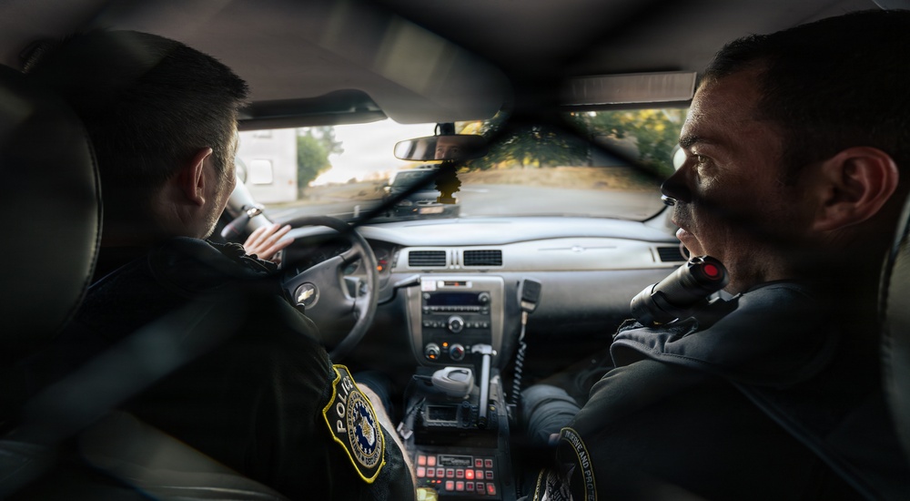 Two uniformed police officers are seated inside a patrol vehicle, viewed from the rear seat. The officer in the driver's seat is facing forward while the passenger officer is turned slightly, holding what appears to be a radio or communication device. The interior shows standard law enforcement vehicle equipment including a console with radio equipment and controls.