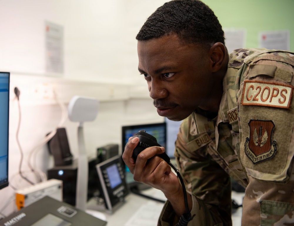 A U.S. Air Force service member wearing OCP (Operational Camouflage Pattern) uniform is leaning over a workstation and speaking into a radio handset microphone. The uniform displays a 'C2OPS' patch and what appears to be a 'U.S. Air Forces in Europe' shield patch. The setting appears to be a command and control operations center with multiple computer monitors and communications equipment.