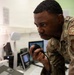 A U.S. Air Force service member wearing OCP (Operational Camouflage Pattern) uniform is leaning over a workstation and speaking into a radio handset microphone. The uniform displays a 'C2OPS' patch and what appears to be a 'U.S. Air Forces in Europe' shield patch. The setting appears to be a command and control operations center with multiple computer monitors and communications equipment.