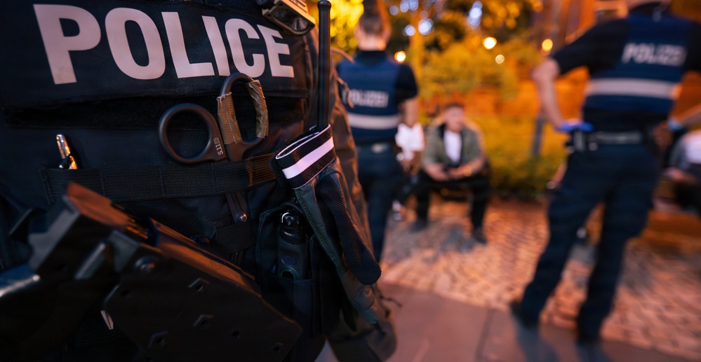 A nighttime law enforcement scene showing multiple police officers in tactical gear on what appears to be a cobblestone street or plaza. The foreground features a close-up of a police vest with visible equipment including scissors, a radio, and other tactical tools. In the background, officers appear to be interacting with or detaining individuals.
