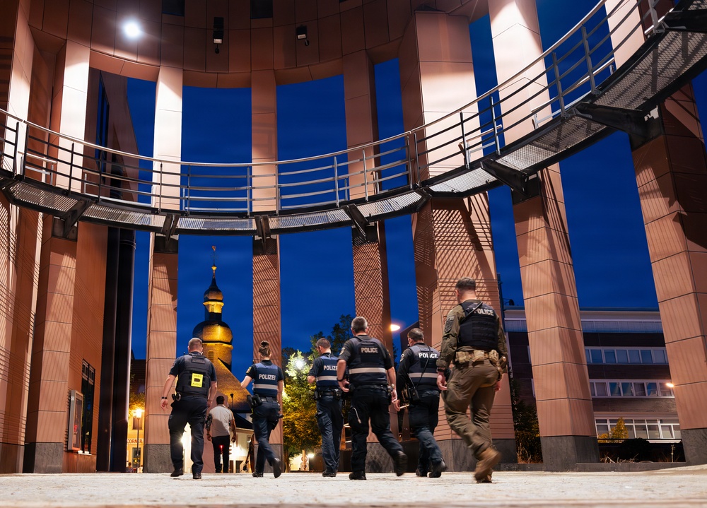 A group of law enforcement officers, including both German 'Polizei' and 'Police'-labeled personnel, are walking together through a modern circular architectural structure at night. The scene appears to depict a joint patrol or security operation in an urban setting. A historic church tower with a golden dome is visible in the background, suggesting a German city center.