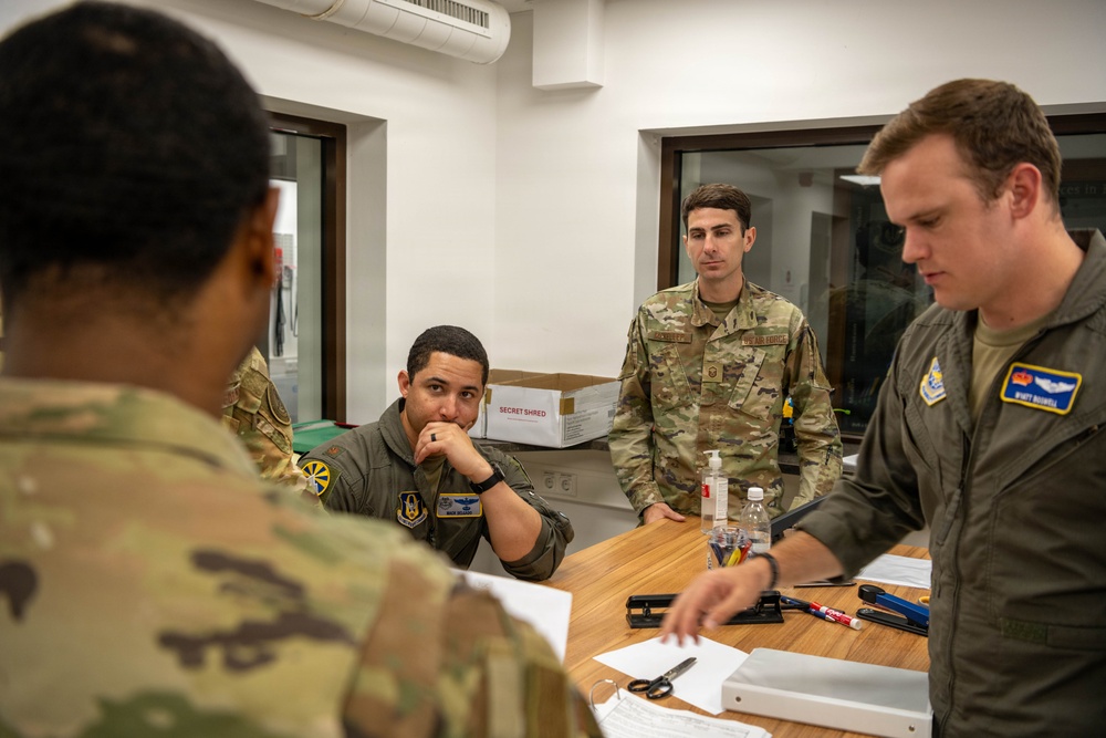 Several U.S. Air Force personnel are gathered around a wooden table in what appears to be an administrative or operations room. Two individuals are wearing olive green flight suits with unit patches, while others wear OCP (Operational Camouflage Pattern) uniforms. They appear to be reviewing documents or conducting a briefing.