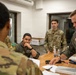 Several U.S. Air Force personnel are gathered around a wooden table in what appears to be an administrative or operations room. Two individuals are wearing olive green flight suits with unit patches, while others wear OCP (Operational Camouflage Pattern) uniforms. They appear to be reviewing documents or conducting a briefing.