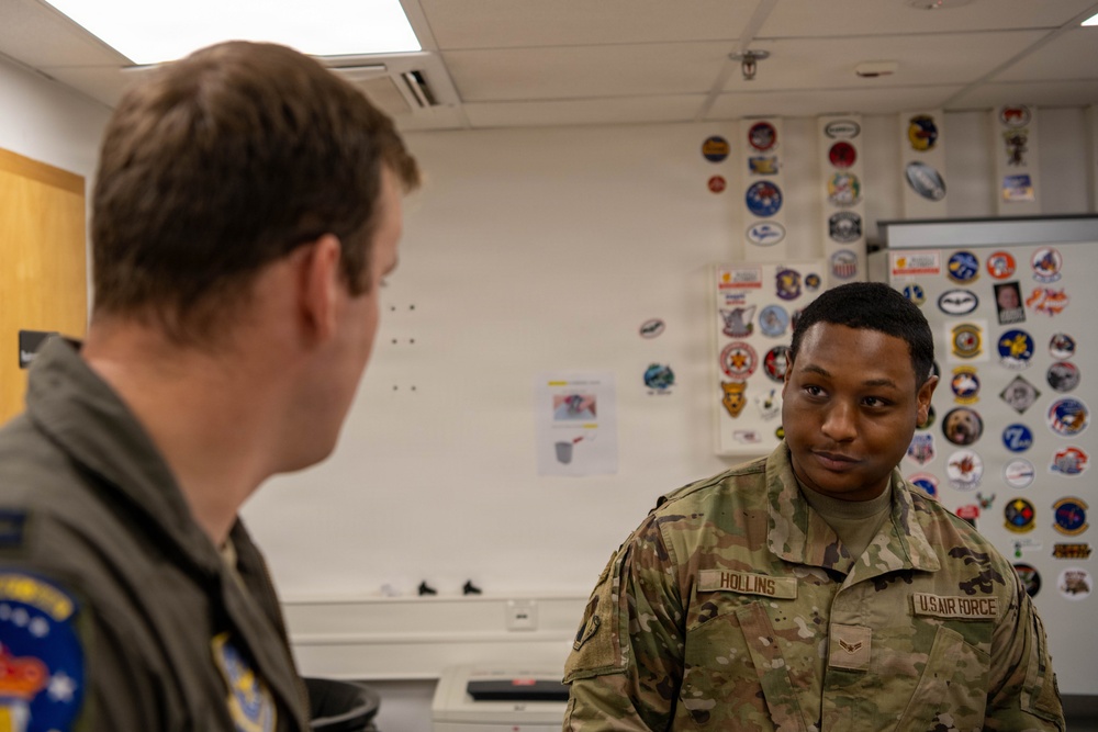 Two U.S. Air Force personnel are engaged in conversation inside what appears to be a military briefing room or office. The airman facing the camera wears an OCP (Operational Camouflage Pattern) uniform with the name tape 'HOLLINS' and 'U.S. AIR FORCE' clearly visible. The background features a whiteboard covered with numerous military unit patches, stickers, and insignia.