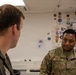 Two U.S. Air Force personnel are engaged in conversation inside what appears to be a military briefing room or office. The airman facing the camera wears an OCP (Operational Camouflage Pattern) uniform with the name tape 'HOLLINS' and 'U.S. AIR FORCE' clearly visible. The background features a whiteboard covered with numerous military unit patches, stickers, and insignia.
