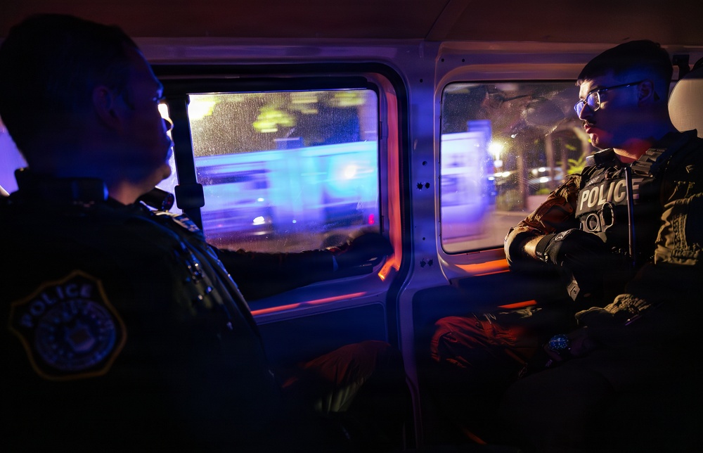 Two police officers are seated inside what appears to be a police transport vehicle or van at night. Both officers are wearing tactical gear with visible 'POLICE' markings, and the scene is illuminated by blue and red emergency lights visible through the rain-speckled windows. The motion blur outside suggests the vehicle may be in transit or surrounded by active emergency response activity.