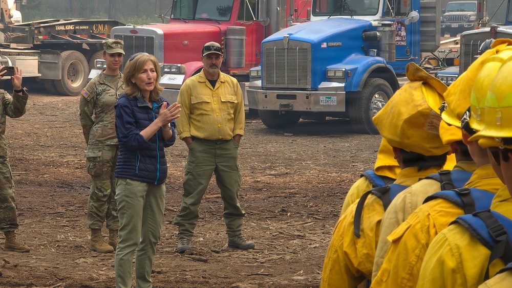 A woman in civilian clothing appears to be briefing or addressing a group of wildfire firefighters in yellow protective gear and hard hats at what appears to be a fire staging area. She is accompanied by a military soldier in camouflage uniform and a man in a yellow fire-resistant shirt. Multiple large heavy-duty trucks are visible in the background, consistent with a wildfire incident command or staging location.