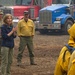 A woman in civilian clothing appears to be briefing or addressing a group of wildfire firefighters in yellow protective gear and hard hats at what appears to be a fire staging area. She is accompanied by a military soldier in camouflage uniform and a man in a yellow fire-resistant shirt. Multiple large heavy-duty trucks are visible in the background, consistent with a wildfire incident command or staging location.