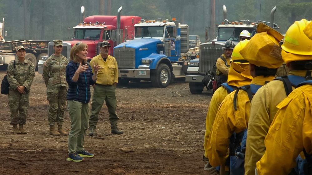 A civilian woman in a blue jacket and green pants appears to be addressing a group of wildland firefighters in yellow Nomex gear and hard hats at what appears to be a fire staging area. Military personnel in camouflage uniforms and a man in a yellow fire shirt stand nearby. Large semi-trucks are visible in the background amid smoky, hazy conditions.
