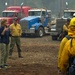 A civilian woman in a blue jacket and green pants appears to be addressing a group of wildland firefighters in yellow Nomex gear and hard hats at what appears to be a fire staging area. Military personnel in camouflage uniforms and a man in a yellow fire shirt stand nearby. Large semi-trucks are visible in the background amid smoky, hazy conditions.