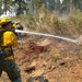 Wildland firefighters in standard yellow Nomex gear are working to suppress or mop up a forest fire in a coniferous woodland setting. The foreground firefighter is directing a pressurized water stream from a red hose onto scorched ground, while additional crew members are visible in the background. The terrain shows significant burn damage with charred soil, ash, and partially burned vegetation.