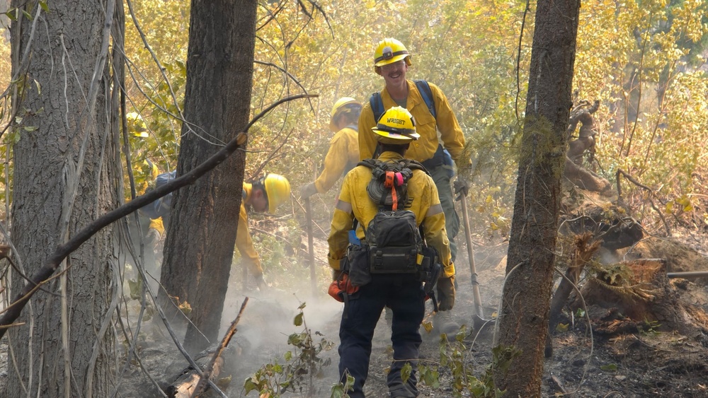 A crew of wildland firefighters in yellow Nomex protective gear and hard hats work through a smoky, partially burned forest. At least four crew members are visible navigating through charred undergrowth and smoke. One firefighter's helmet is labeled 'WRIGHT' and the crew appears to be conducting mop-up or fire line operations.