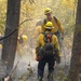 A crew of wildland firefighters in yellow Nomex protective gear and hard hats work through a smoky, partially burned forest. At least four crew members are visible navigating through charred undergrowth and smoke. One firefighter's helmet is labeled 'WRIGHT' and the crew appears to be conducting mop-up or fire line operations.