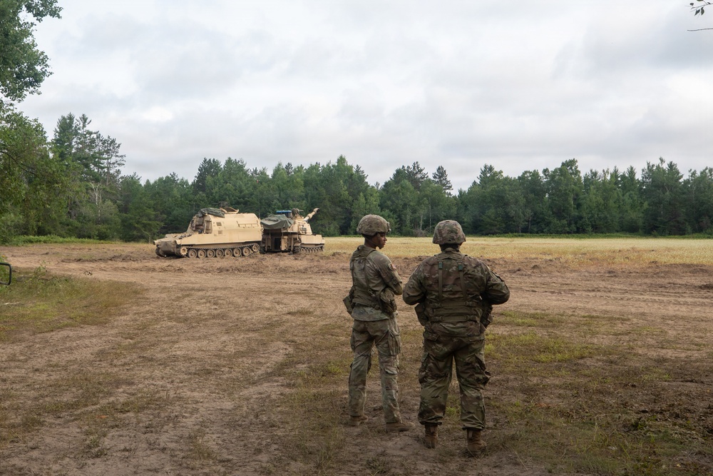 Two soldiers in camouflage uniforms and helmets stand in a dirt field observing a tracked military vehicle, likely an M109 Paladin self-propelled howitzer or similar armored artillery platform. The scene appears to be a training exercise at a military installation, with a treeline of mixed conifers and deciduous trees forming the background. The overcast sky and muddy terrain suggest recent or ongoing field operations.